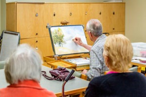 Two women watching an elderly man paint